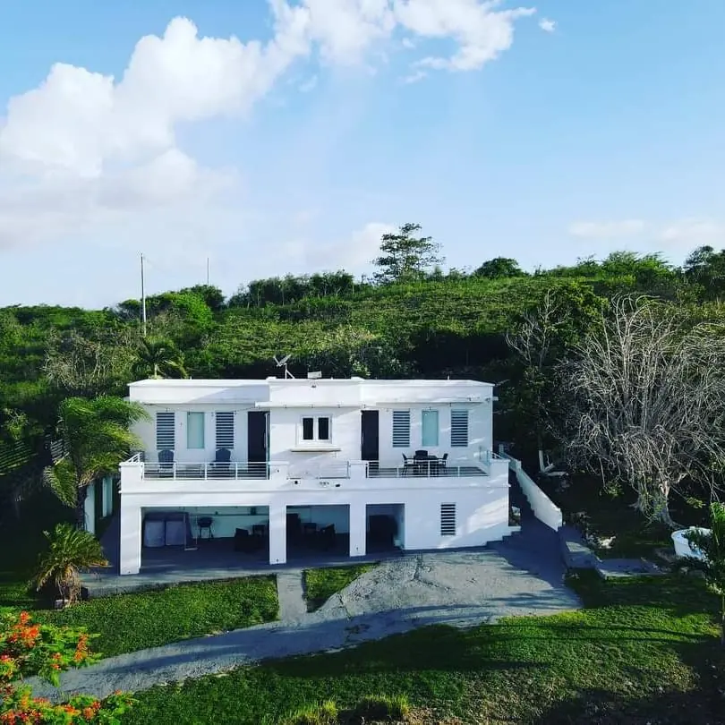 The front of the house, white with grey balcony railings, on a green hill under a blue sky